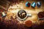 Tazza di caffè vista dall’alto con mappa del mondo sulla superficie, circondata da chicchi, capsule, cialde e polvere: rappresentazione del confronto tra caffè in grani e in capsule.